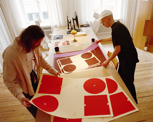 Two men standing on either side of a long table, both flipping through large art prints with the text "OH NO" in red and burgundy.