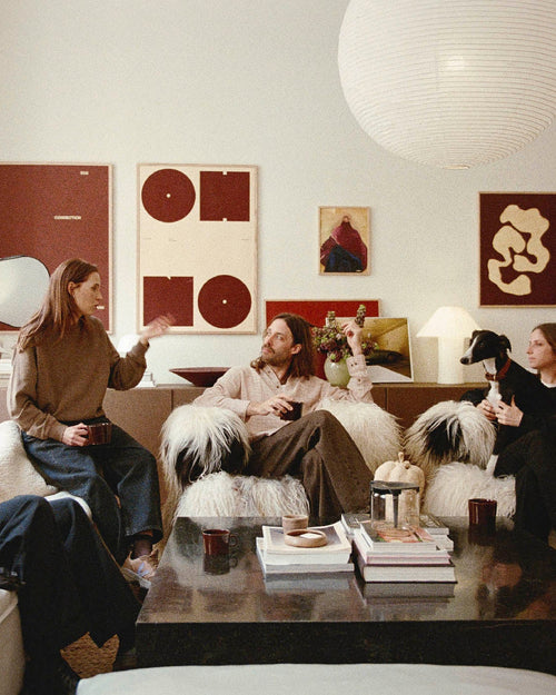 Four people sitting in a living room decorated with large art prints and large paper pendant lamps, chatting. Two on a couch to the left, two in large furry chairs straight ahead, one with a whippet-like dog in her lap. Woman on couch is speaking to the man and woman in the chair.