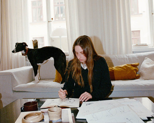 Woman (artist Anna Mörner) sitting on floor in front of a large grey couch, sketching in sketchbook. A whippet-like dog standing on the couch in the background.