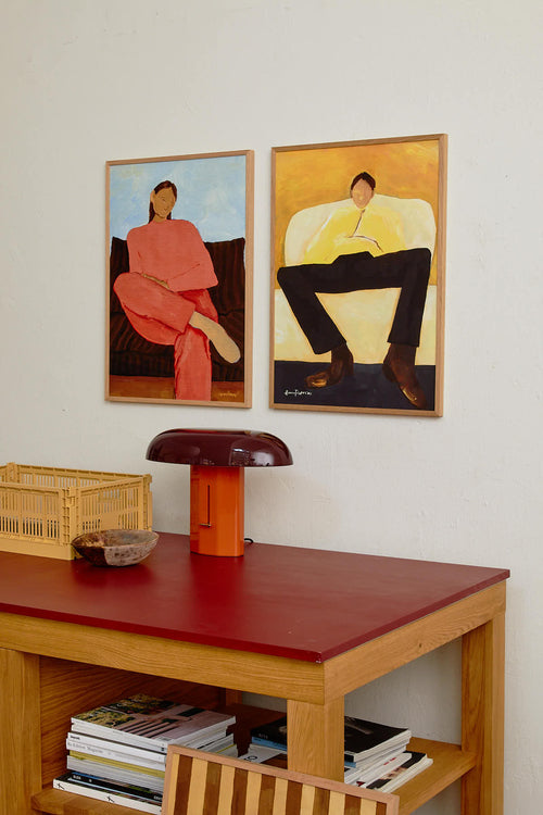 Two framed paintings of people on a wall above a wooden table with a red surface.