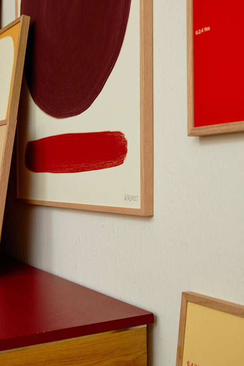 Framed abstract art pieces on a wall with a red shelf below.
