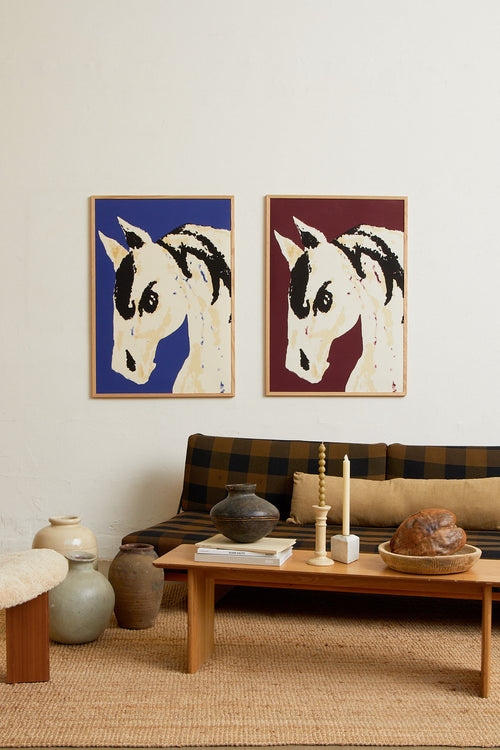 Art print with a stylized illustration of a horse head in light beige and black tones against a cobalt blue background, with an intentionally blurred and painterly feel. Styled in a minimalist interior.