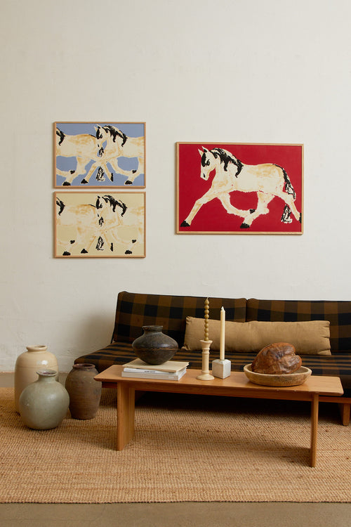 Art print with a stylized illustration of two mirrored horses in motion in light beige and black tones against a yellow-beige background, with an intentionally blurred and painterly feel. Styled in a minimalist interior.
