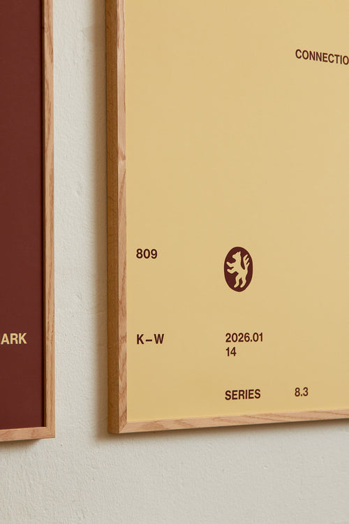 Art print with minimalist, small graphic text elements and a symbol motif in brown against a beige background. Styled in a minimalist interior.