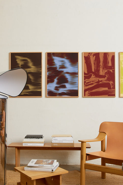 Art print featuring soft, blurred contours and flowing light blue shapes emerging against a dark red-brown surface, creating an impression of motion and impermanence. Styled in a minimalist interior.