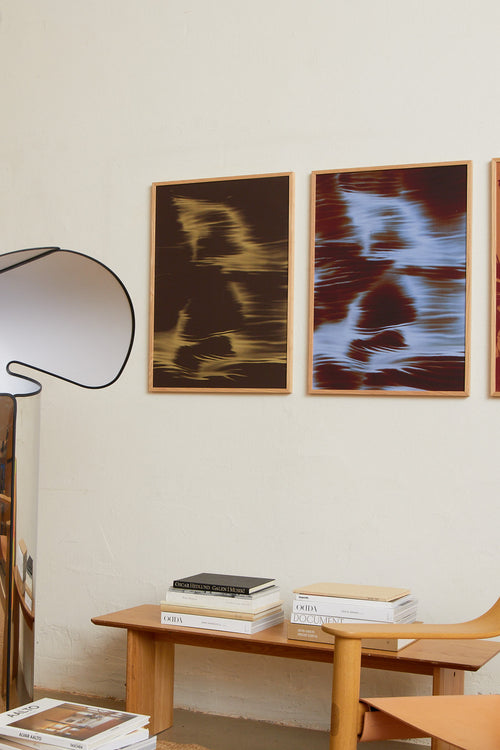 Art print featuring soft, blurred contours and flowing light blue shapes emerging against a dark red-brown surface, creating an impression of motion and impermanence. Styled in a minimalist interior.