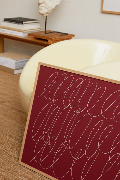 Art print with a graphic illustration of sweeping, continuous lines in a light tone against a red background, with a minimalist and abstract expression. Styled in a minimalist interior.