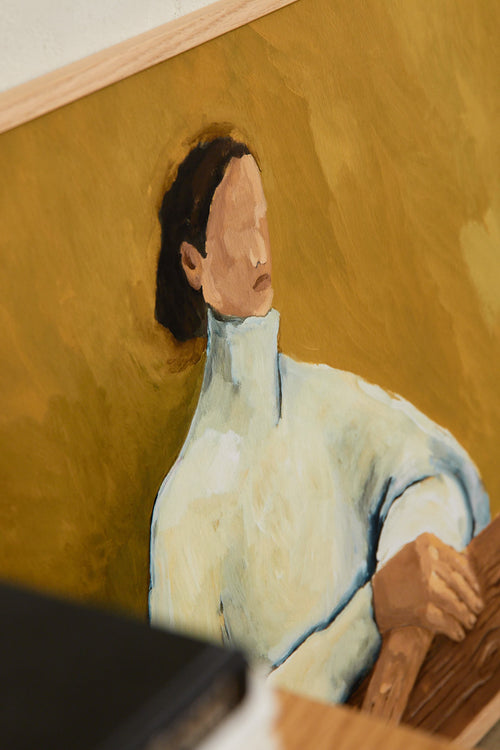 Art print featuring a painting of a person in a white chunky turtleneck against an olive-colored background, resting their hands on a wooden bench. Styled in a minimalist interior.