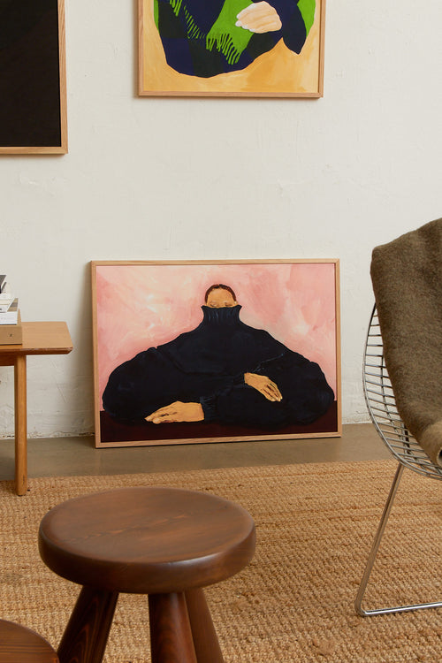 Art print featuring a figurative painting of a seated person in an oversized dark sweater, with the face partially hidden, against a light pink background. Styled in a minimalist interior.