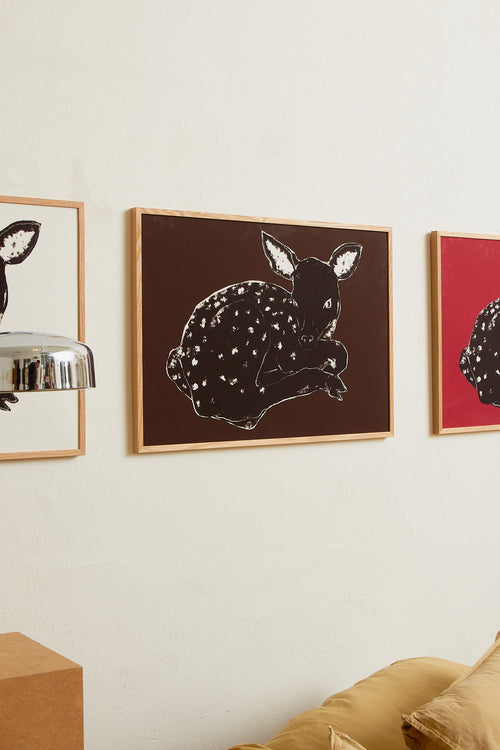 Art print with a stylized illustration of a lying deer in a dark tone against a dark brown background, with a minimalist and graphic expression. Styled in a minimalist interior.