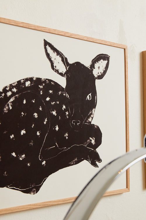 Art print with a stylized illustration of a lying deer in a dark tone against a, with a minimalist and graphic expression. Styled in a minimalist interior.