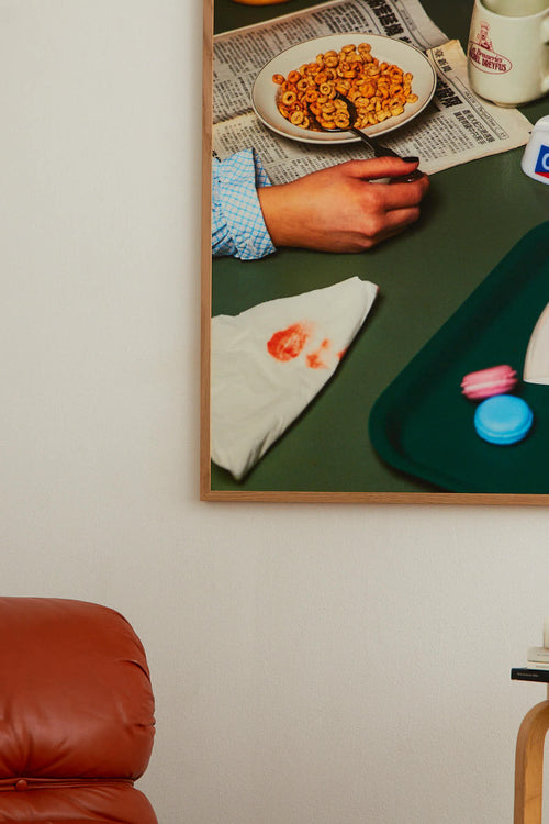 Framed artwork of a person eating cereal with a green tray and objects on a white wall.