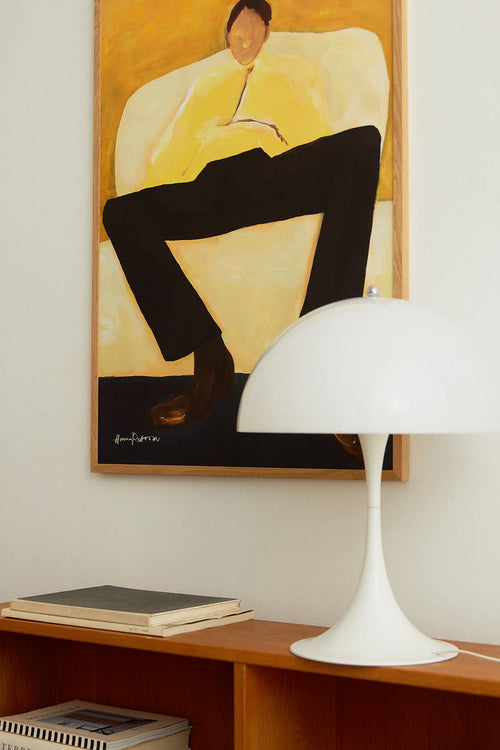 White lamp on a wooden shelf with a framed painting of a person in a yellow chair on the wall.