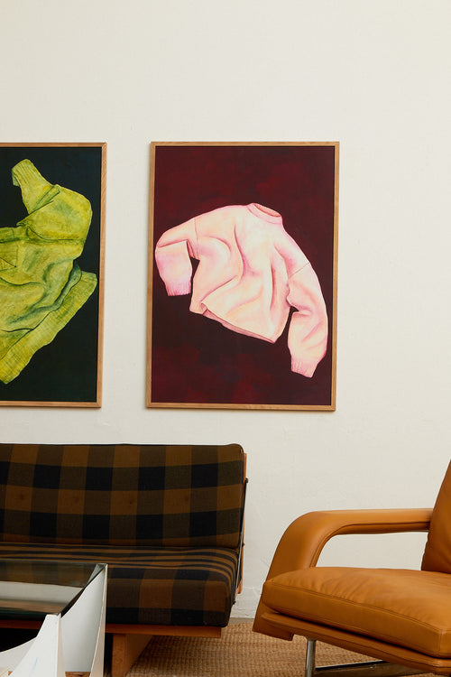 Two framed artworks of green and pink garments on a wall with a checkered sofa and brown chair.