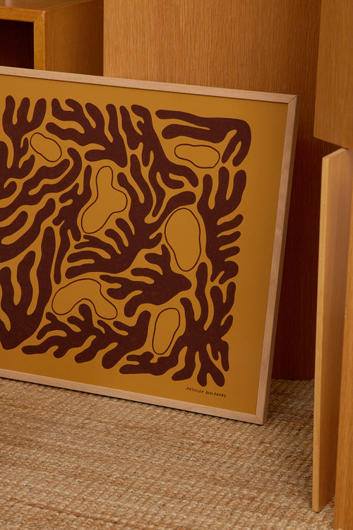 Framed abstract artwork with brown patterns on a yellow background, leaning against a wooden wall.