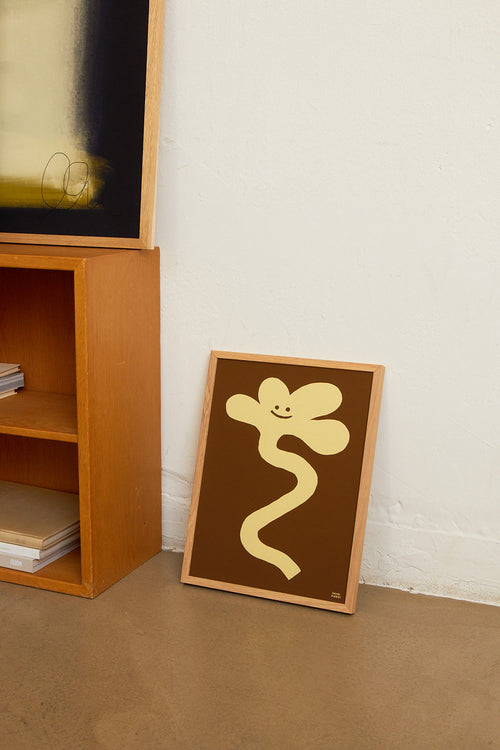 Framed artwork with a yellow abstract design on a brown background, leaning against a white wall next to a wooden shelf.