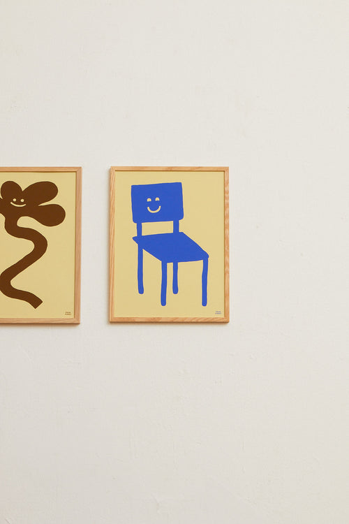 Two framed artworks on a wall, one with a blue chair and smiley face, the other with a brown abstract figure.