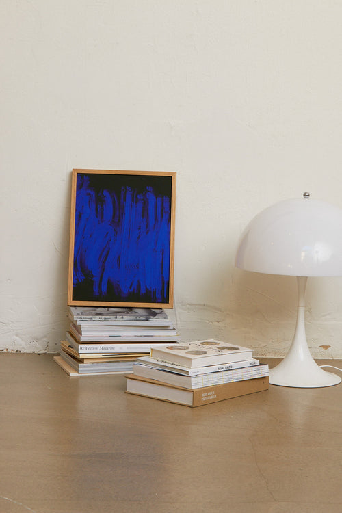 Stack of books with a framed blue artwork and a white lamp on a wooden floor.