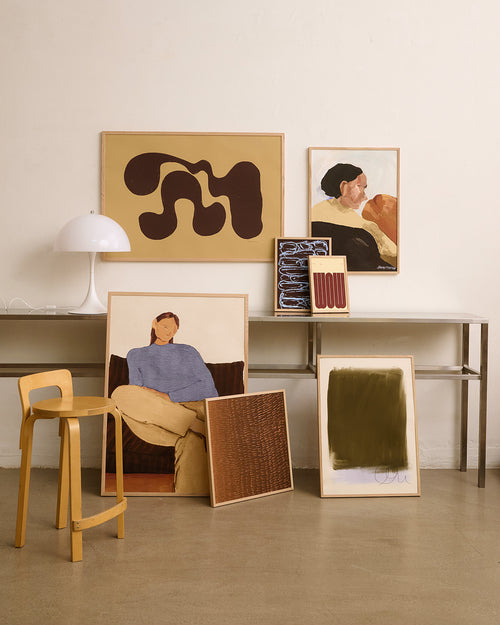 A styled interior scene with multiple framed artworks leaning and hanging against a cream wall above a metal console table. The artworks include abstract shapes, portraits, and textured abstract compositions in neutral and earthy tones.