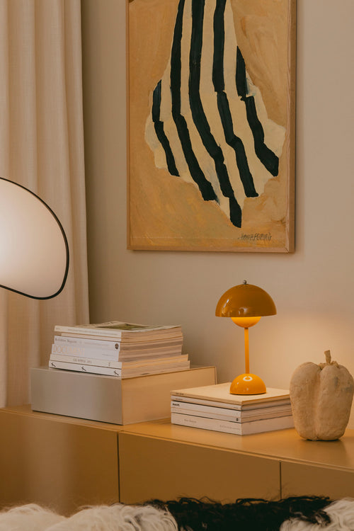 Decorative shelf with books, a yellow lamp, and a textured object against a neutral wall with abstract art.